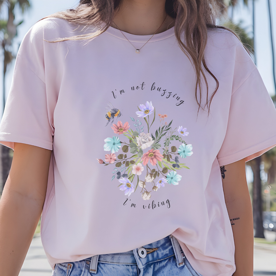 Close-up view of the "I'm Not Buzzing" graphic, showcasing the detailed illustration of the bee with its delicate wings and fuzzy body, the intricate details of the wildflower petals and leaves, and the whimsical typography of the "I'm Not Buzzing" and "I'm Vibing" text. Color Light pink