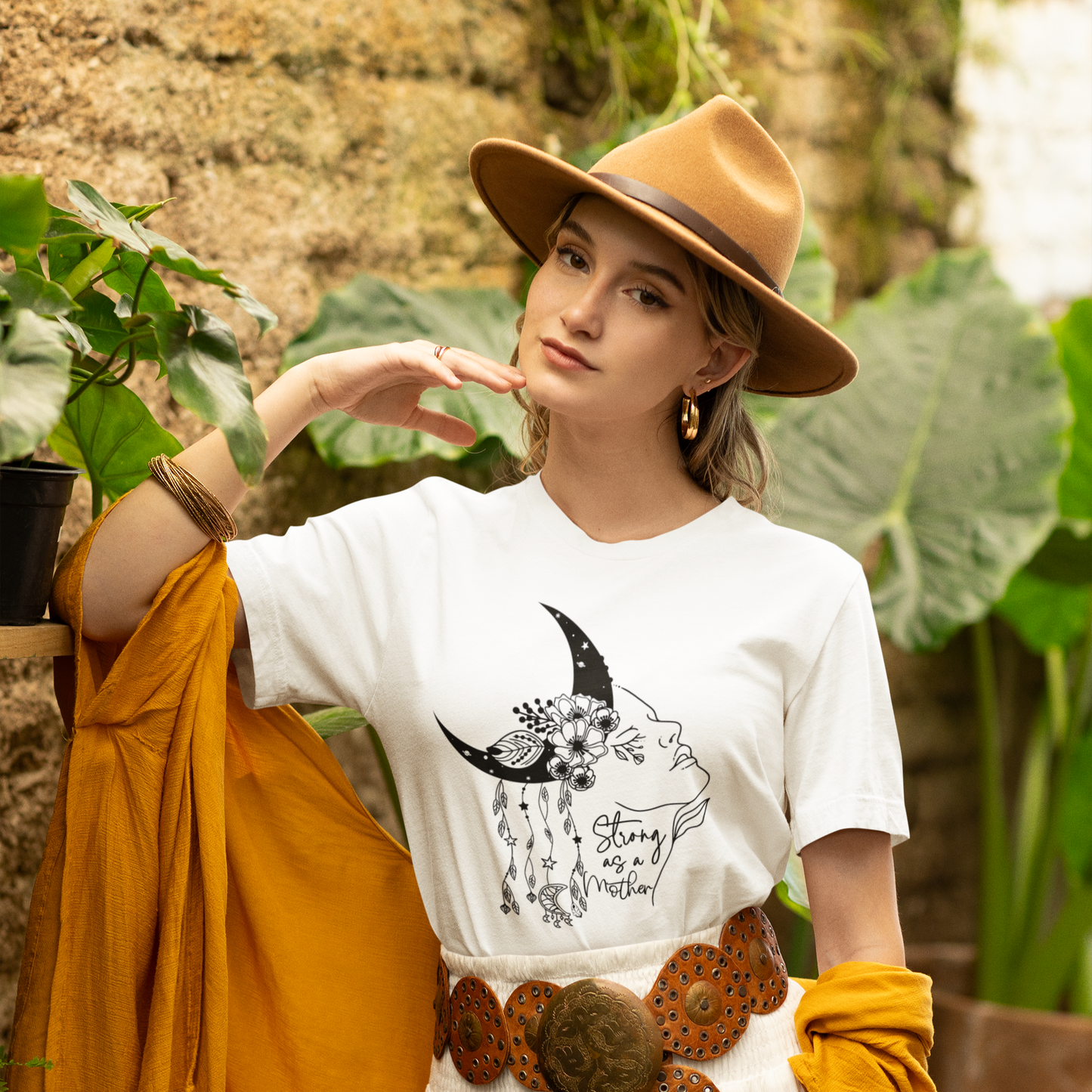 "Strong as a Mother" Unisex Jersey T-Shirt, featuring a powerful graphic of a woman's profile emerging from a crescent moon, adorned with blooming flowers and delicate dangling charms, modeled by a Woman  wearing size M.