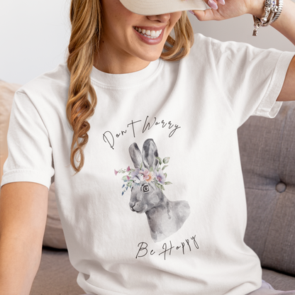 "Don't Worry, Be Hoppy!" Unisex Garment-Dyed T-Shirt, featuring a charming watercolor illustration of a grey rabbit wearing a crown of delicate pink and purple flowers, with the text "Don't Worry Be Hoppy" in a playful script above and below, modeled by a woman wearing size L. Color White