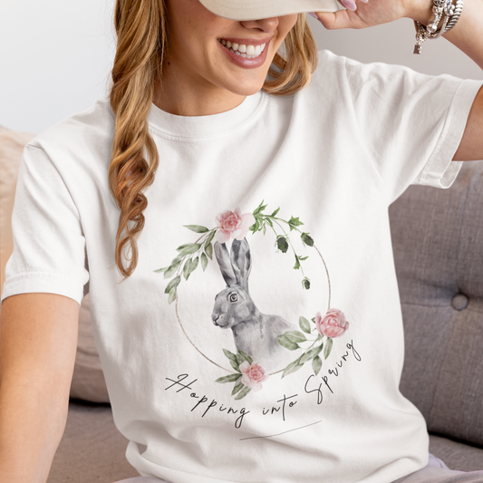 "Hoppin' into Spring" Unisex Garment-Dyed T-Shirt, featuring a delicate watercolor illustration of a grey rabbit framed by a circular wreath of pink roses and lush green foliage, modeled by a Woman wearing size L (oversized). Color White